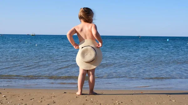 かわいい女の子3歳のわらの帽子のダンスと海を見て、バックビュー、クローズアップ。子供時代、関係、夏休みの概念。帽子に焦点を当て. — ストック写真