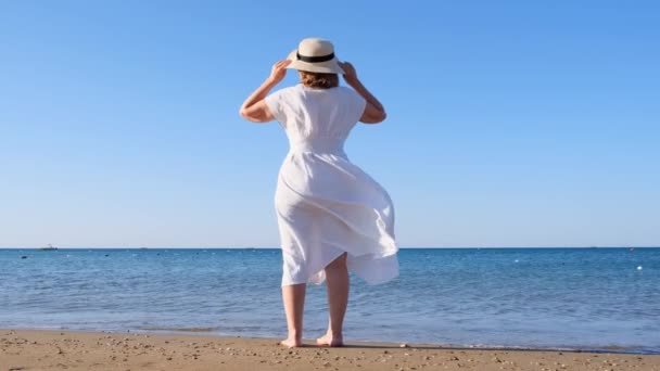 Задний вид на зрелую женщину в соломенной шляпе и белом платье прогулки вдоль синего побережья моря в солнечный летний день, наслаждаясь свободой и отдыхом. Концепция типичной жизни пенсионеров. — стоковое видео
