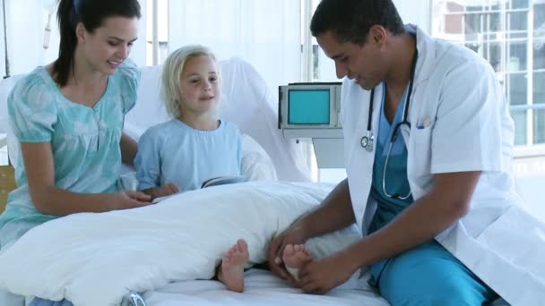 Médico vendando el pie de una pequeña paciente acostada en la cama con su madre — Vídeo de stock