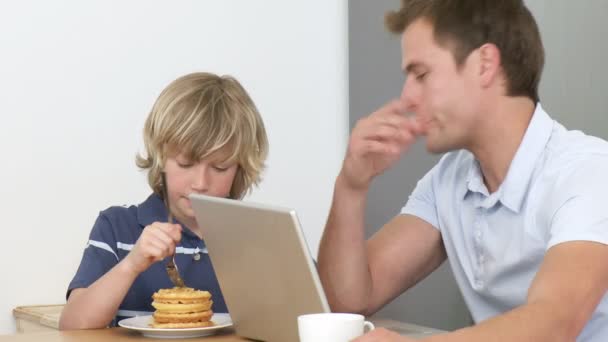 Vater mit Laptop und Sohn essen ein Süßes in der Küche — Stockvideo