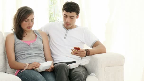Casal de adolescentes comendo hambúrgueres e conversando no sofá — Vídeo de Stock