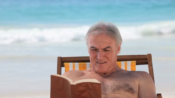 Starszy mężczyzna siedzi na krześle plaży książką — Wideo stockowe