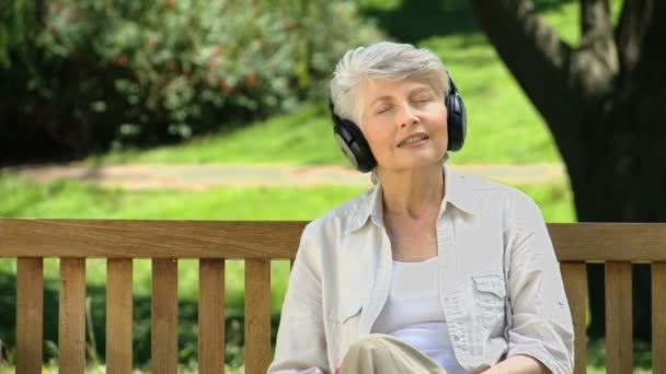 Mujer vieja escuchando música con auriculares en un banco — Vídeos de Stock