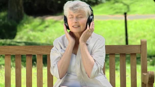 Starší žena poslechu hudby s headsetem na lavičce — Stock video