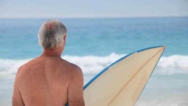 Elderly man looking at the ocean with a surfboard — Stock Video