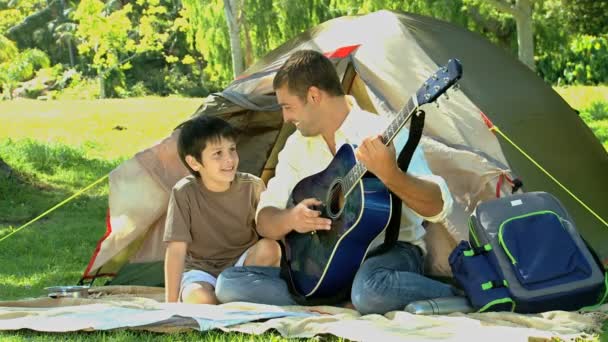 Padre tocando la guitarra a su hijo delante de una tienda de campaña — Vídeo de stock