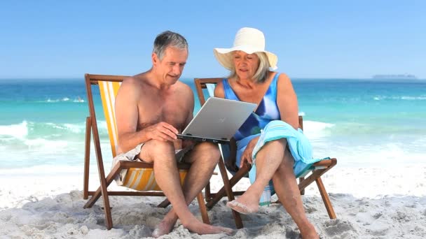 Bejaarde echtpaar met behulp van een laptop zitting op strandstoelen — Stockvideo