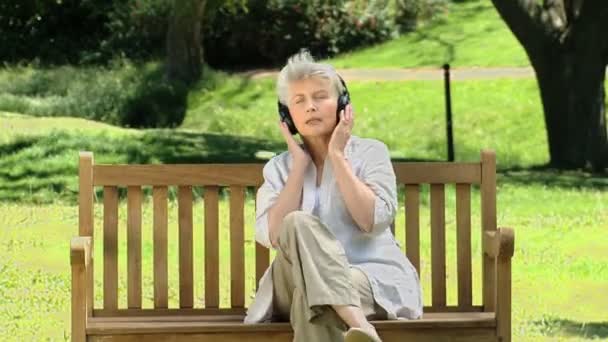 Anciana escuchando música con auriculares en un banco — Vídeo de stock