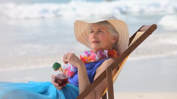 Oudere vrouw op zoek naar de horizon met een cocktail zittend op een strandstoel — Stockvideo