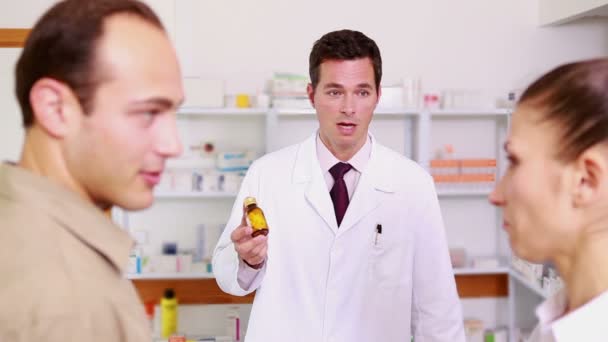 Улыбающийся фармацевт указывает на фляжку с таблетками мужчине и женщине — стоковое видео