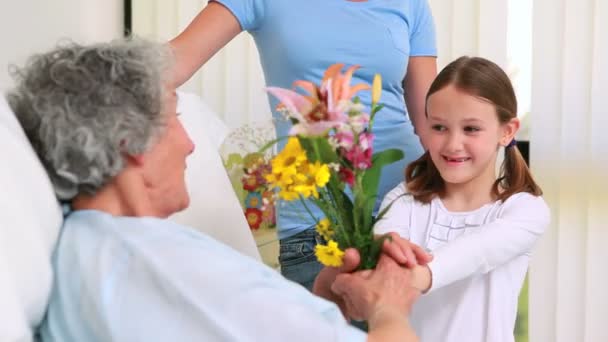 Mujer sonriente y niña ofreciendo flores a una anciana en una cama de hospital — Vídeo de stock