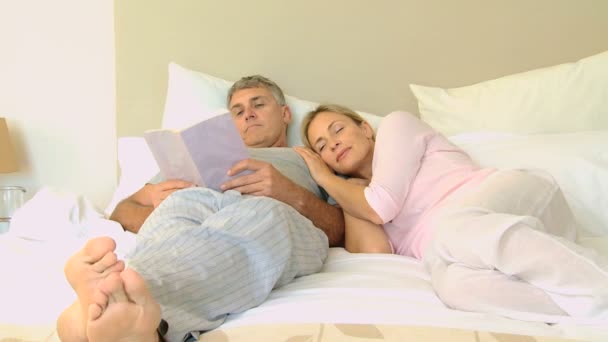 Young wife on bed snuggling up to husband reading a book — Stock Video