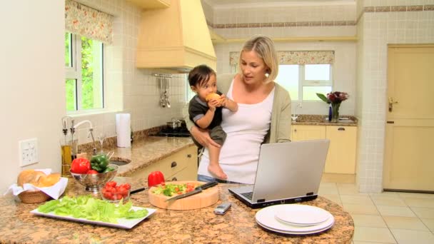 Young mother in kitchen working on laptop holding her baby — Stock Video