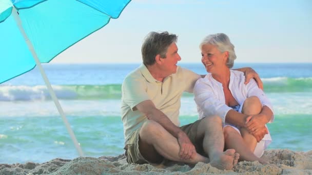 Elderly couple laughing under a beach umbrella — Stock Video