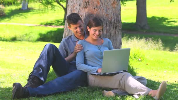 Jeune couple avec un ordinateur portable appuyé contre un arbre — Video