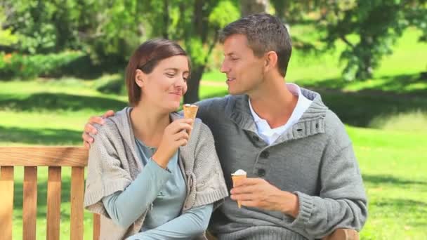 Pareja joven comiendo helado en un banco del parque — Vídeo de stock