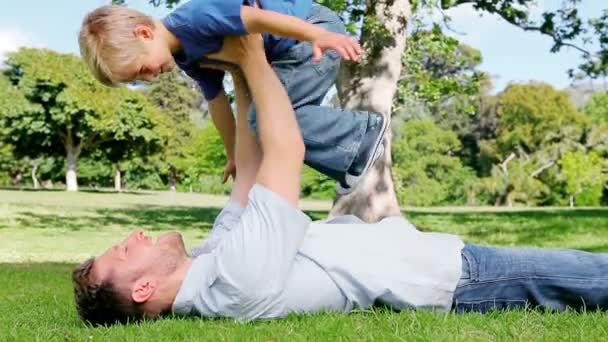 A father lifts his son above him as he lies on the grass before returning him to the ground — Stock Video