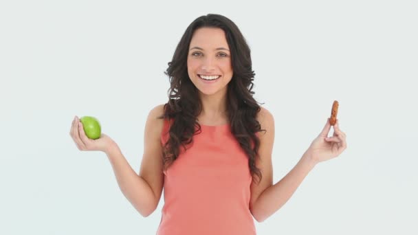 Молодая привлекательная женщина выбирает между яблоком и печеньем — стоковое видео