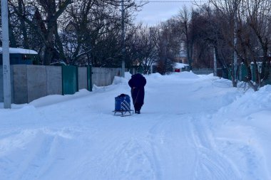 Resimde, kırsal bir yol boyunca karla kaplı bir kızağın üzerinde ağır bir yük taşıyan bir kadın görülüyor..