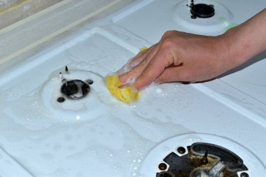 Resimde, bir kadın gaz sobasının yüzeyini sünger ve deterjanla yıkıyor..