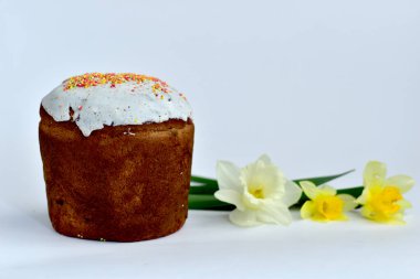 Beyaz bir çiçekle süslenmiş Paskalya pastası ve beyaz bir masanın üzerinde bir buket nergis var..