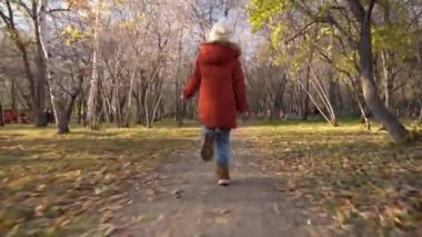 Küçük, mutlu, altı yaşında, şapkalı ve ceketli bir kız, Ekim ormanında turuncu yapraklarla koşar ve ellerini sallar. Parkta veya meydanda sonbahar yürüyüşü. Bir çocuğun tasasız çocukluğu..
