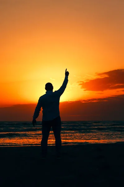 a silhouette of a man pointing towards the sky during sunset at the seaside.