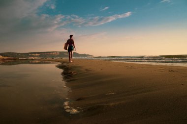 Genç sporcu gün doğumunda plajda kaykayını tutuyor.