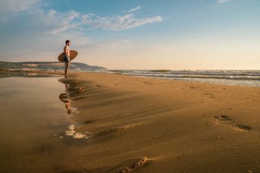 Genç sporcu gün doğumunda plajda kaykayını tutuyor.