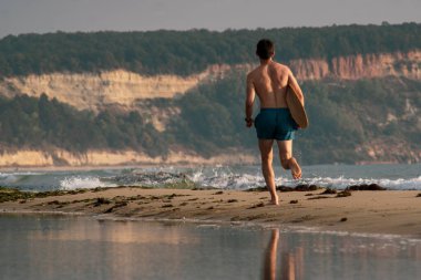Genç sporcu adam gün doğumunda plajda kaykayıyla koşuyor.