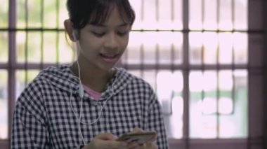 Sıradan elbiseli Asyalı güzel kız kulaklık takıyor ve evindeki cep telefonundan arkadaşlarıyla konuşuyor. Neşeli genç kız, akıllı telefonuyla ailesiyle sohbet ediyor..