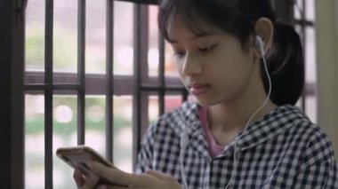 Tayland etnik kökenli sevimli genç kız, evde pencerenin yanında dururken akıllı telefonuyla arkadaşlarıyla sohbet etmeyi sever. Güzel genç kız cep telefonundan mesaj atıyor..