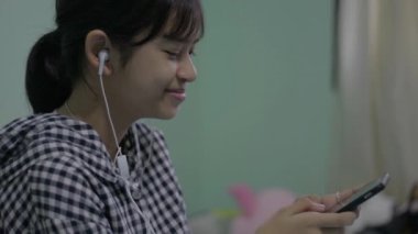 Asyalı mutlu bayan genç kız evde kulaklıklarla arkadaşlarıyla sohbet ederken eğleniyor ve gülüyor. Ortaokul öğrencisi cep telefonuyla sosyal iletişim kuruyor.