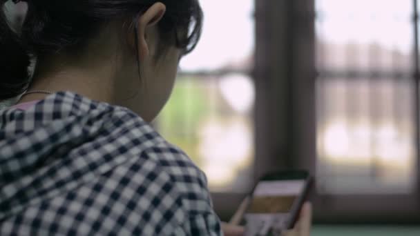 自宅滞在中に携帯電話上のフッテージストックウェブサイトからビデオクリップファイルを視聴し 選択するアジアの女の子の背面ビュー ネットをサーフィン インターネットブラウジング技術 家の隔離 — ストック動画