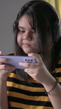 Dikey portre Asyalı tombul yüzlü genç kız evde otururken atıştırmalık yerken akıllı telefonuyla oyun oynamaktan hoşlanıyor. Mutlu, şişman, genç kadın yaşam tarzını kapat..