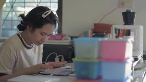 作業机の上にデジタルタブレットを入力カジュアルなドレスでアジアのかなり若い女性 自宅で勉強中にデジタルデバイス上の友人にオンライン女子大生のテキストメッセージ — ストック動画