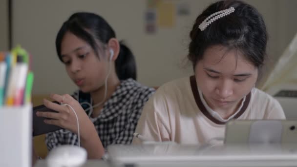 亚洲漂亮的女大学生喜欢在桌上摆弄数字设备 而她的妹妹则喜欢在家里用耳机看智能手机上的社交视频 手足结连在一起 — 图库视频影像