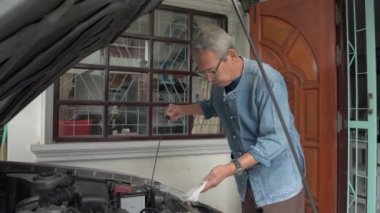Asyalı yaşlı gri saçlı büyükbaba evinin önündeki eski arabasının motor yağı ölçüsünü kontrol ediyor. Boş vakti olan emekli kıdemli bir adam. Sağlıklı yaşlı insan yaşam tarzı.