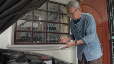 Gözlüklü Asyalı yaşlı büyükbaba evden ayrılmadan önce eski arabasının motor yağı seviyesini kontrol ediyor. Emeklilik hayatı. Araç bakımı.