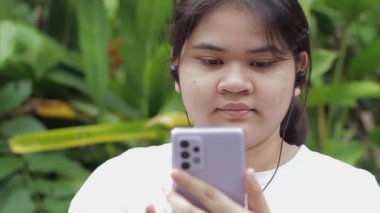 Portre mutlu Asyalı tombul kız yaz sabahı parkta kulaklıklı cep telefonuyla sosyal güvenlik videosu izliyor. Kırsal gençliğin modern hayatı. Genç yaşam tarzı.