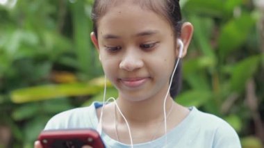 Portre mutlu Asyalı genç kız yaz sabahı parkta kulaklıkla cep telefonuyla sosyal güvenlik videosu izliyor. Kırsal gençliğin modern hayatı. Genç yaşam tarzı.