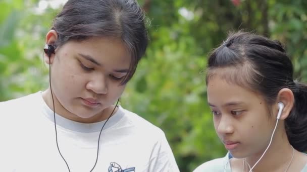 公園内のイヤホン付き携帯電話でオンラインでソーシャルビデオを見ている2人のタイの少女の肖像画 兄弟姉妹の家族との絆 農村部の若者の現代生活 若い生活 — ストック動画