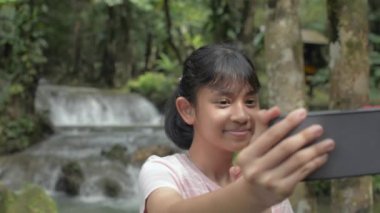 Neşeli Asyalı güzel kız, ormandaki tropik şelalenin yanında akıllı telefonlu bir selfie videosu çekmekten hoşlanıyor. Seyahat tecrübesi. Tayland.