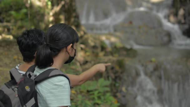 亚洲少女背着背包 戴着面具 喜欢看到美丽的瀑布瀑布在热带雨林的岩石上流淌 徒步旅行丛林探险 — 图库视频影像