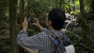 Asyalı bayan sırt çantalı gezgin ormanda yürüyüş yaparken cep telefonuyla fotoğraf çekmeyi ve video kaydetmeyi seviyor. Yazın ormanda tropikal şelale manzarası.