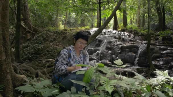 アジアの女性植物研究者は 滝の周りに成長する植物の特性を調べ 森の自然と光合成の生物学的プロセス ノートに書く — ストック動画