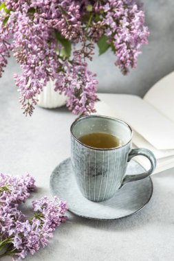 Çiçek açan leylak dalları, çay fincanı ve gri arka plan üzerine açık kitap kompozisyonu