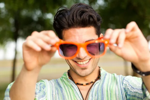 Homem bonito segurando um par de óculos de sol laranja e sorrindo — Fotografia de Stock