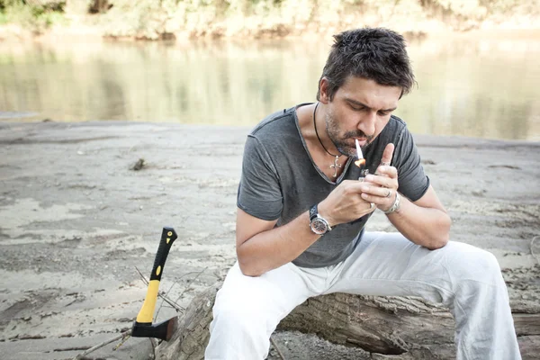 Homem áspero em pé junto ao rio acendendo um cigarro - na série selvagem — Fotografia de Stock