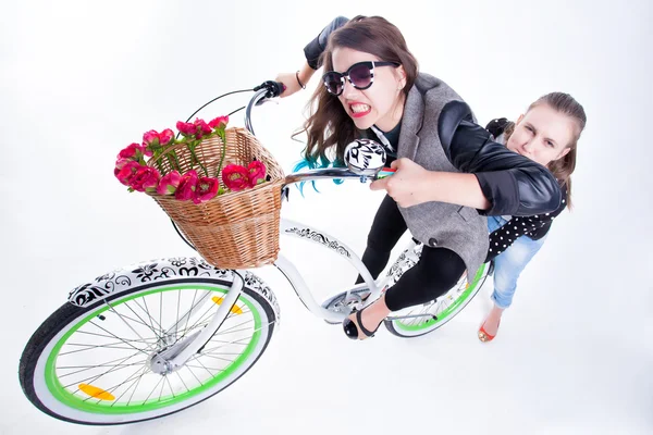 Dos chicas montando en bicicleta haciendo caras graciosas - aisladas sobre fondo azulado — Foto de Stock
